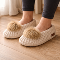 White slippers with brown pom-poms worn indoors on a wooden floor.