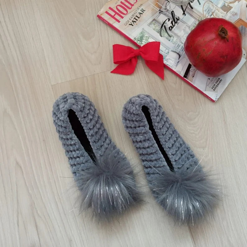 Gray slippers with fluffy pom-poms on a wooden floor with a magazine and pomegranate.