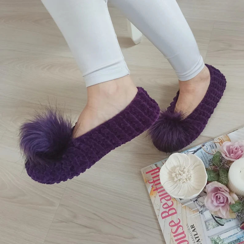 Purple slippers with fluffy pom-poms worn by a person on a wooden floor.