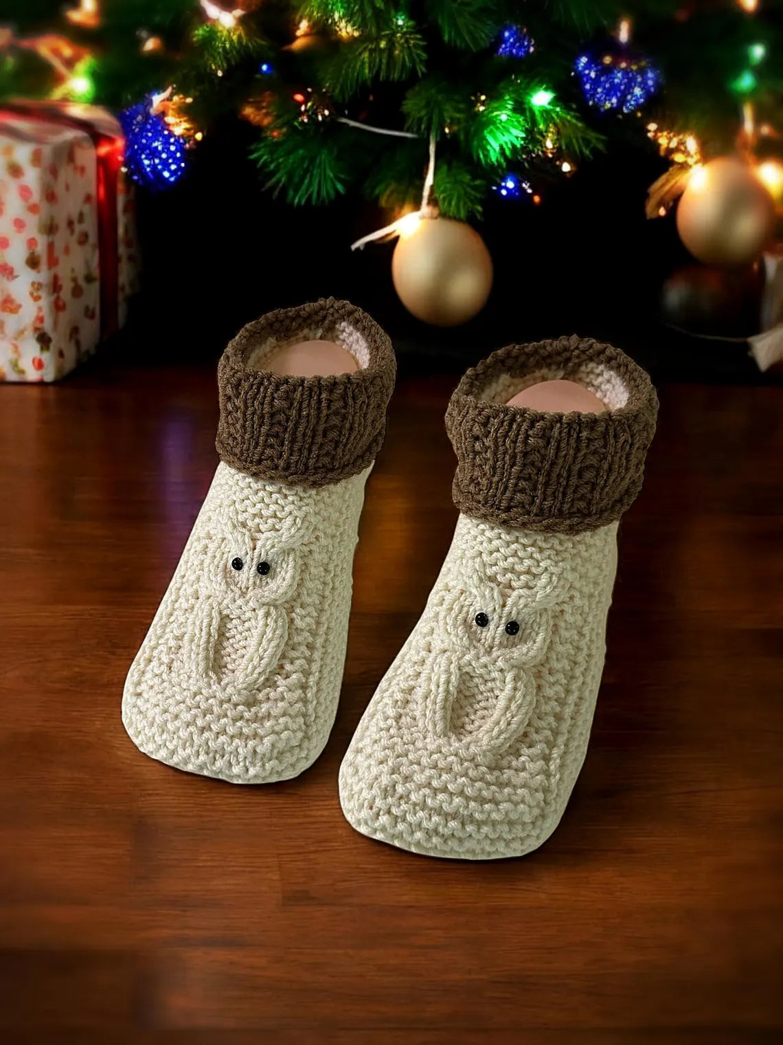 Knitted baby booties with owl design on a wooden surface with Christmas decorations in the background.