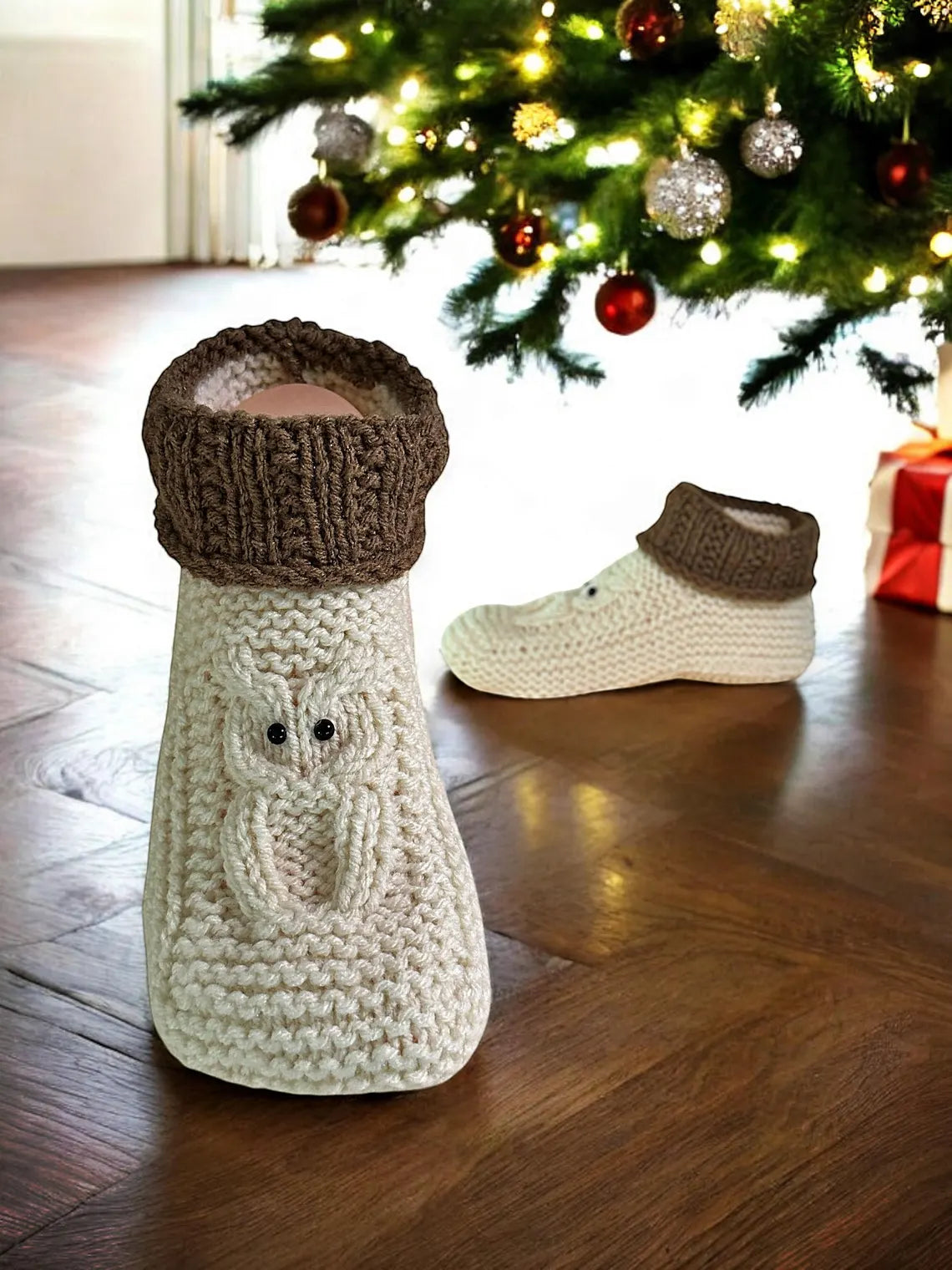 Knitted booties with brown tops on a wooden floor in front of a decorated Christmas tree.