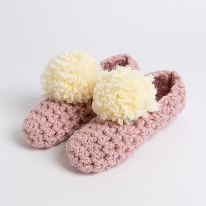 Pink crochet slippers with white pom-poms on a light gray background